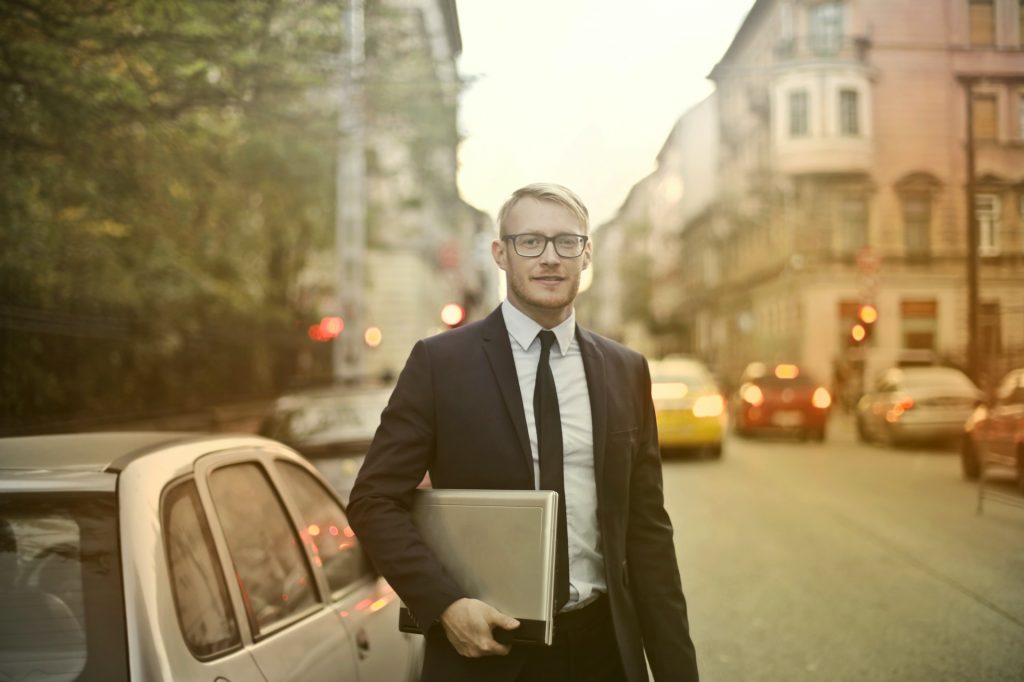 Ein junger Sales Manager geht im schwarzen Anzug mit weißem Hemd zu einem Kundentermin. Er befindet sich in einer Stadt und um ihn herum fahren bzw. parken Autos. Es scheint ein sommerlicher Abend zu sein und der Sales Manager hält einen Laptop unter dem Arm und lächelt in die Kamera.