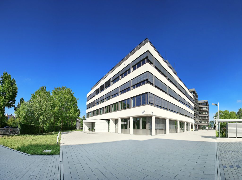 Hier ist der Eingang unserer Firmenzentrale in Nürnberg zu sehen. Das Gebäude ist erst ein paar Jahre alt und verfügt über vier Stockwerke. Die Außenfarbe ist weiß mit großzügigen Fenstern in alle Richtungen.