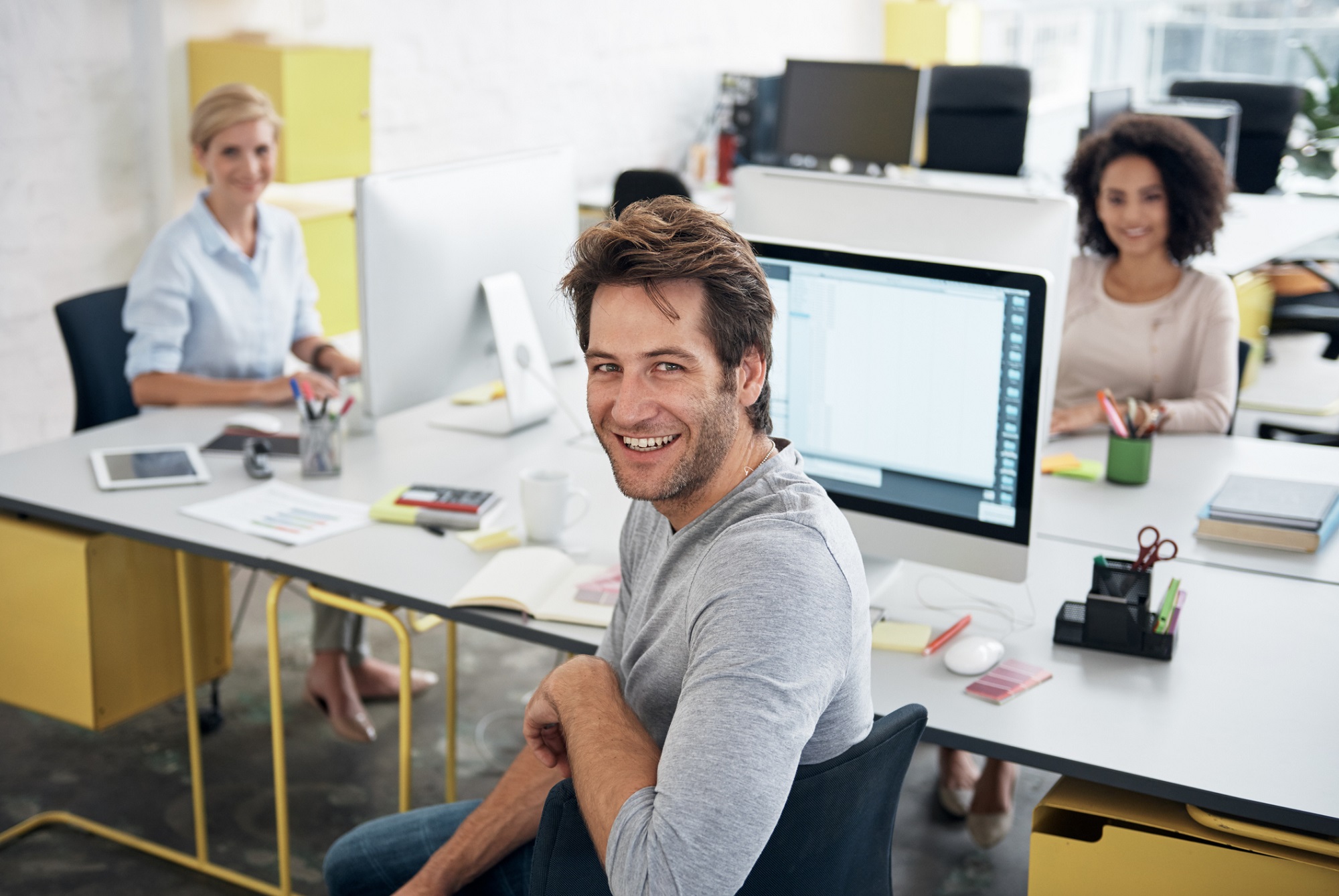 Drei Kollegen in einem modernen Büro: Im Vordergrund ein Mann, der in die Kamera lächelt, im Hintergrund zwei Frauen, die an ihren Schreibtischen arbeiten und ebenfalls lächeln.
